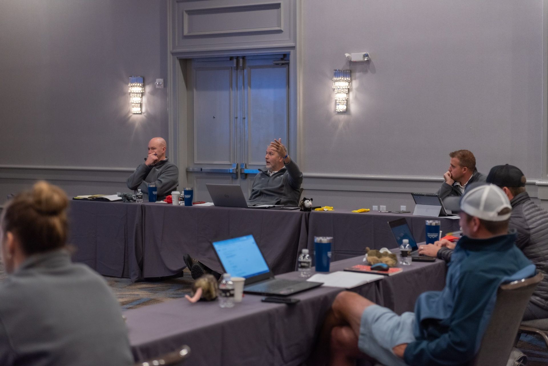 A group of people are sitting at long tables in a conference room.