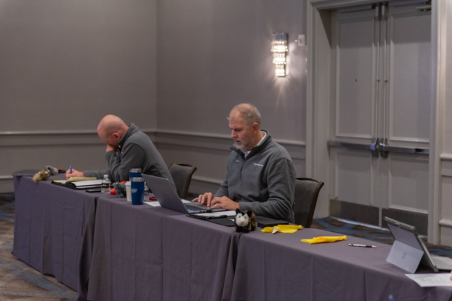 Two men are sitting at a long table with laptops.