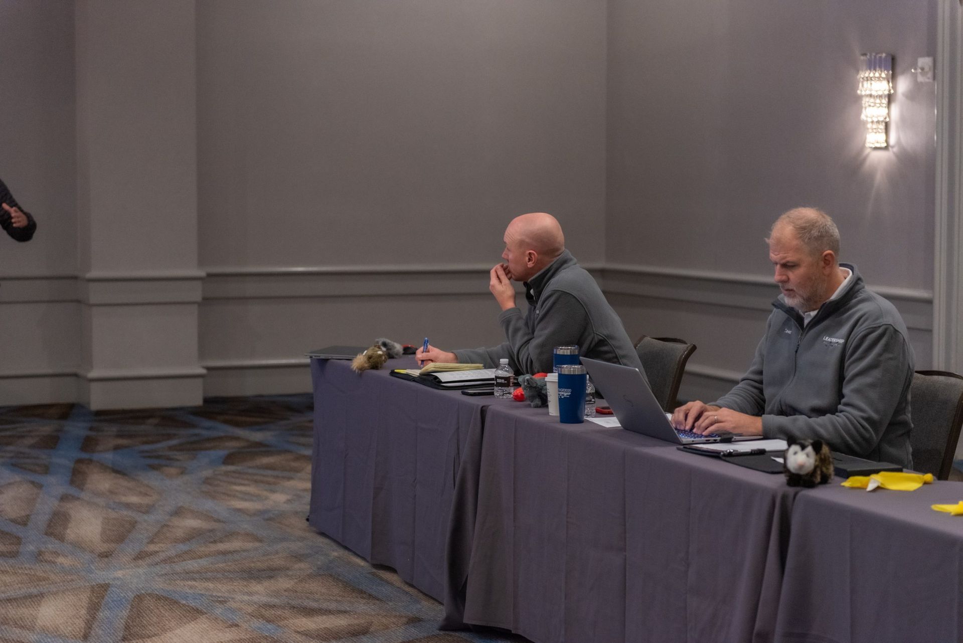 Two men are sitting at long tables with laptops in a conference room.