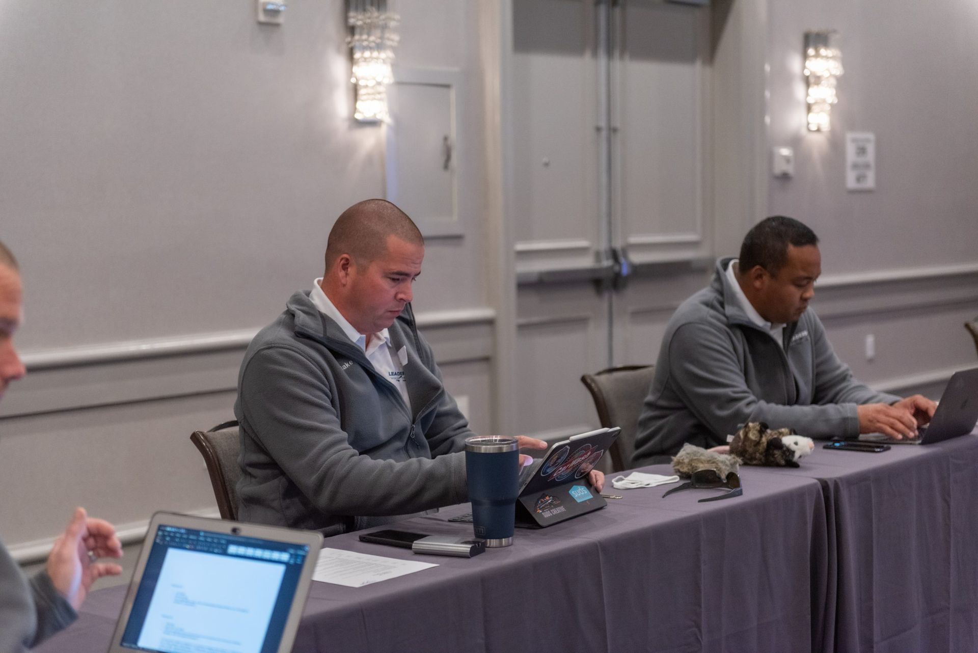 Two men are sitting at a long table with laptops.