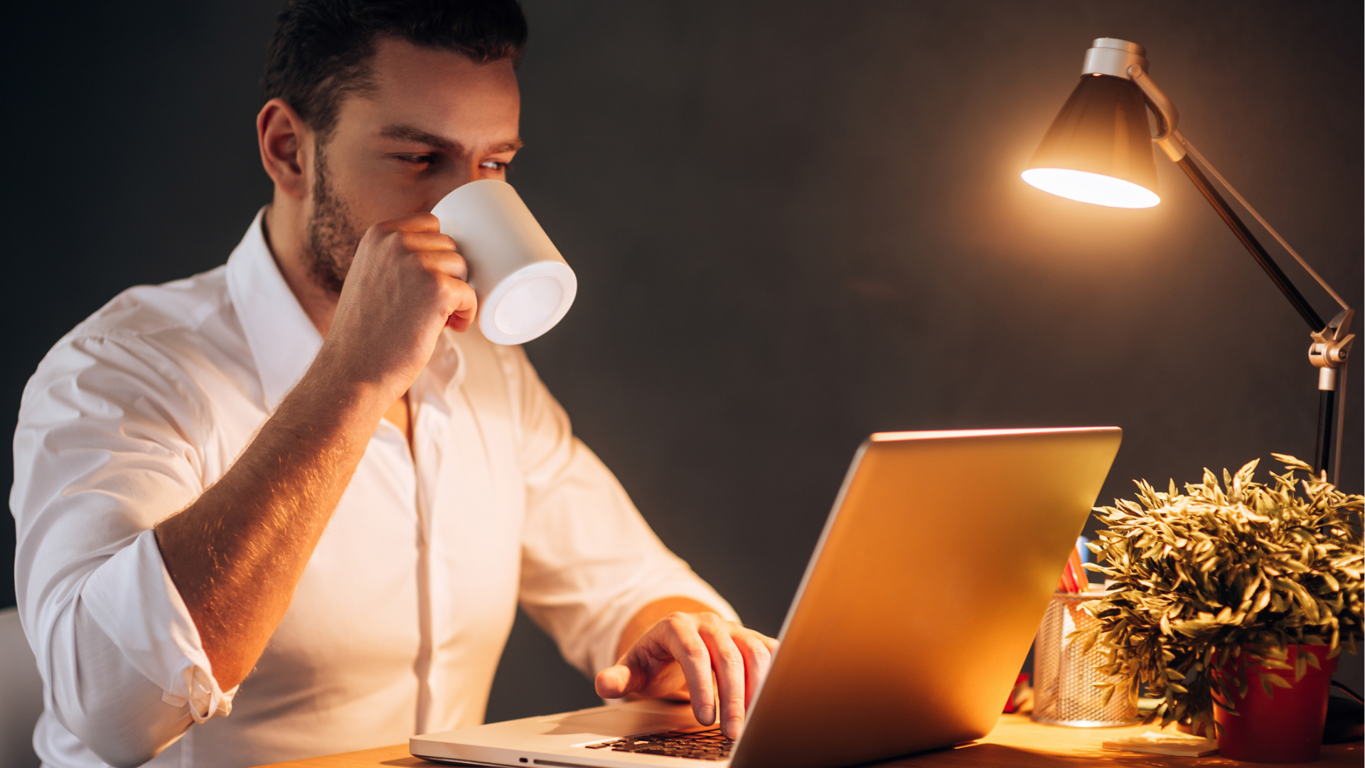 Man with glasses, working on a laptop at a desk with a coffee mug and papers.