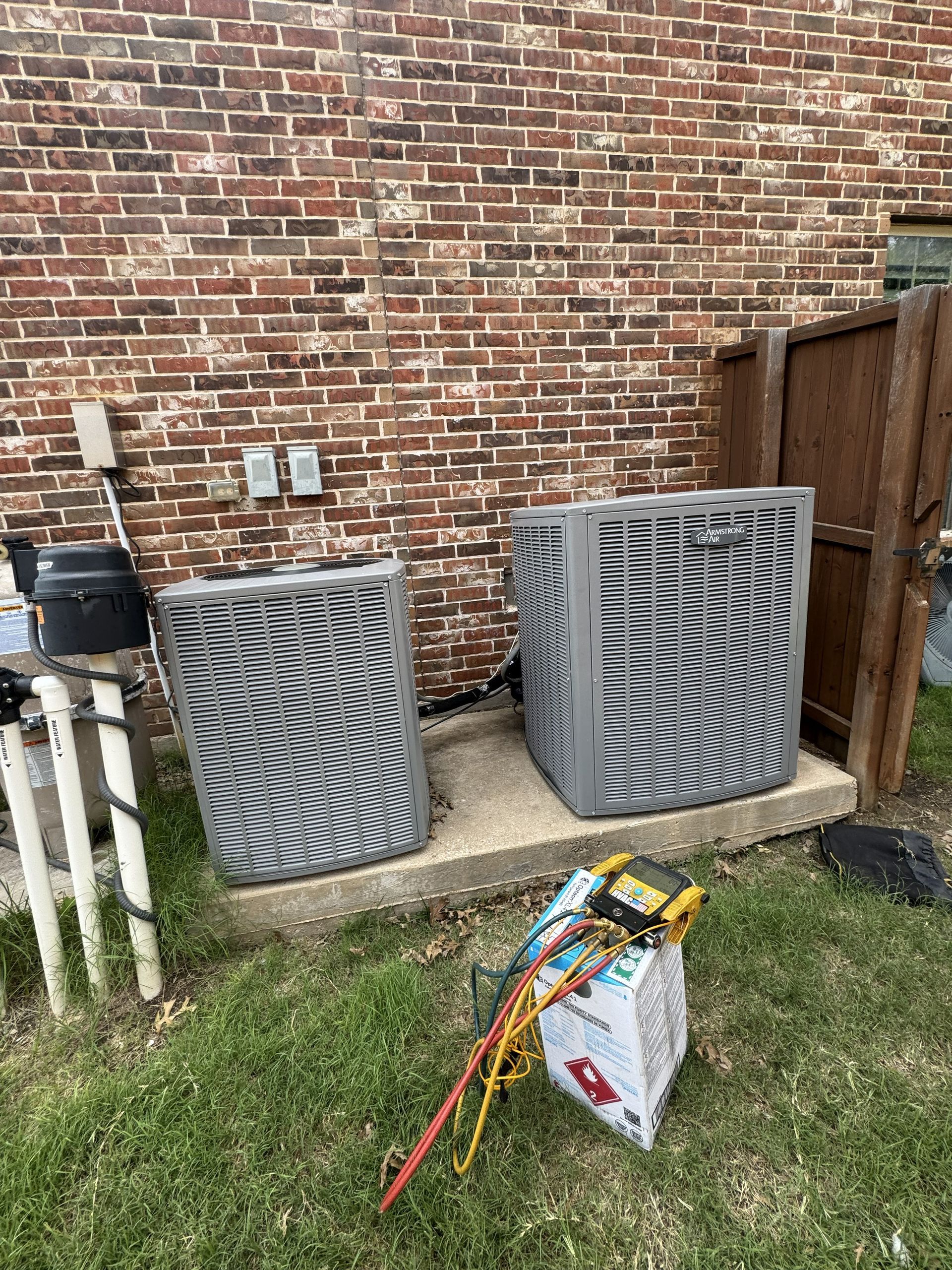 Two air conditioning units on a concrete pad next to a brick wall and a fence. Tools and a box are in front.