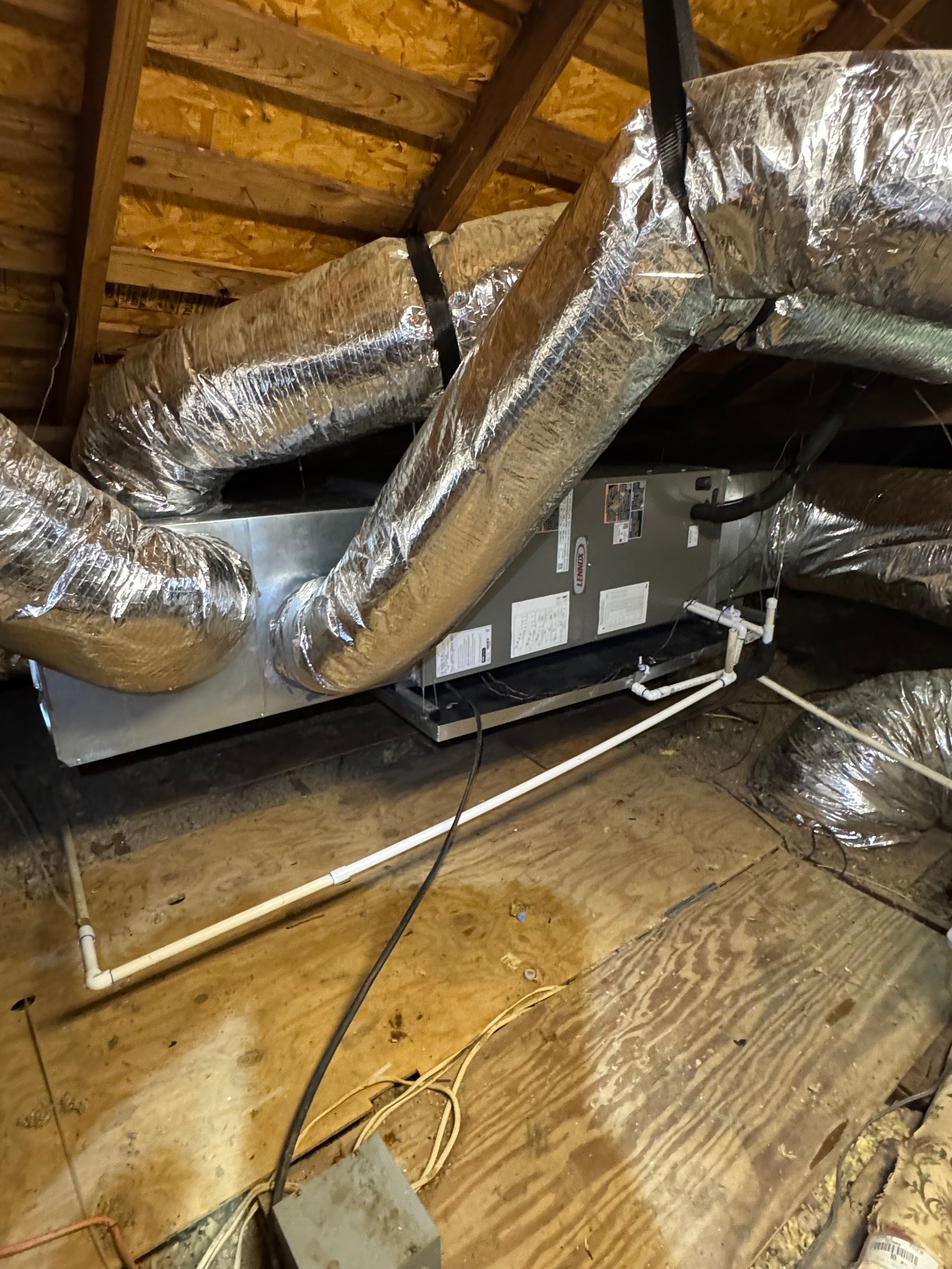 HVAC unit with silver ducts in an attic, sitting on wood. PVC pipe and electrical cables are visible.