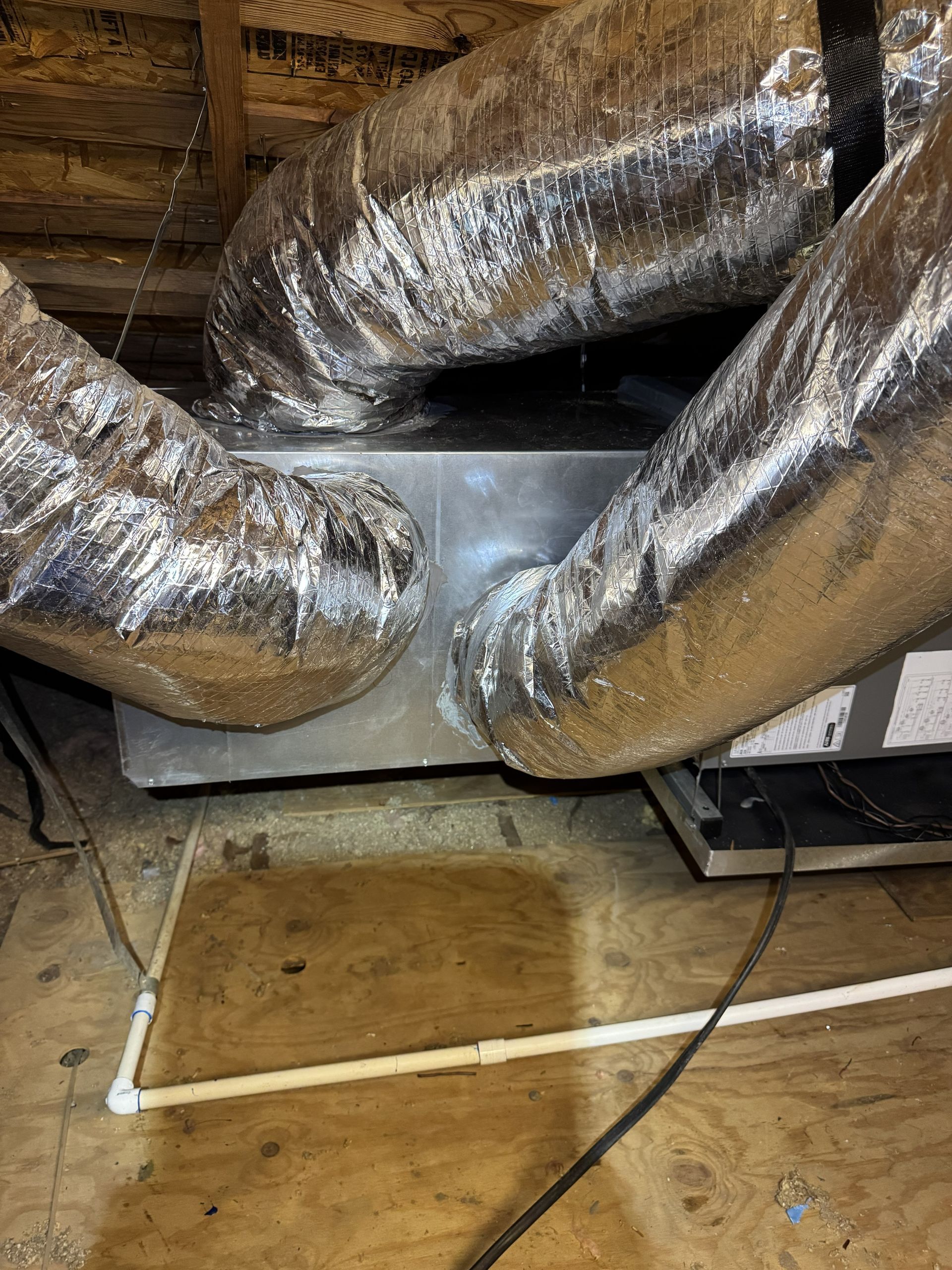 HVAC unit with silver ducts in an attic, sitting on wood. PVC pipe and electrical cables are visible.