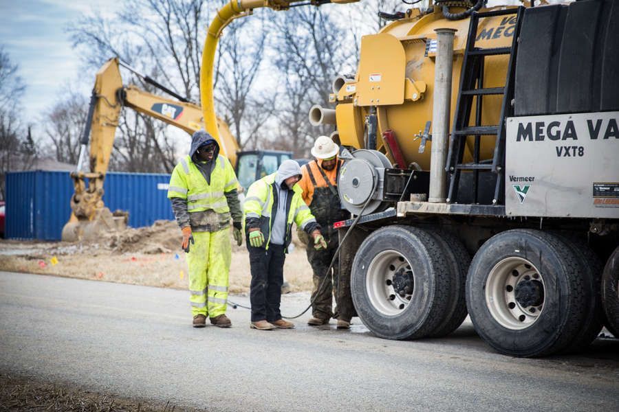 Workers in safety gear near a yellow MEGA VAC truck on a road, with an excavator in the background.