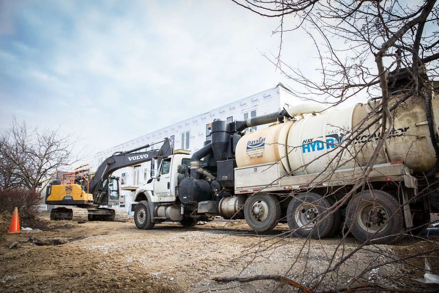 A large white vacuum truck and an excavator are working on a construction site, next to a building under construction.
