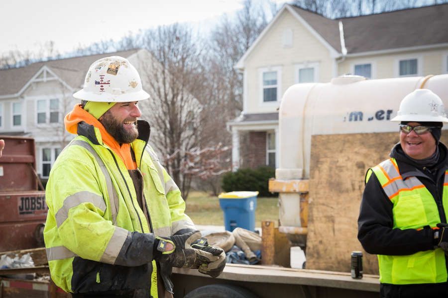 Two construction workers smiling, standing near a water tank and buildings. One wears a yellow jacket, orange hood, and hard hat, while the other wears a safety vest and glasses.