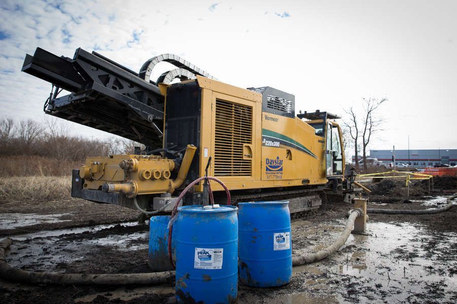 A yellow drill rig with tracks sits in muddy ground, near two blue barrels and a field on an overcast day.