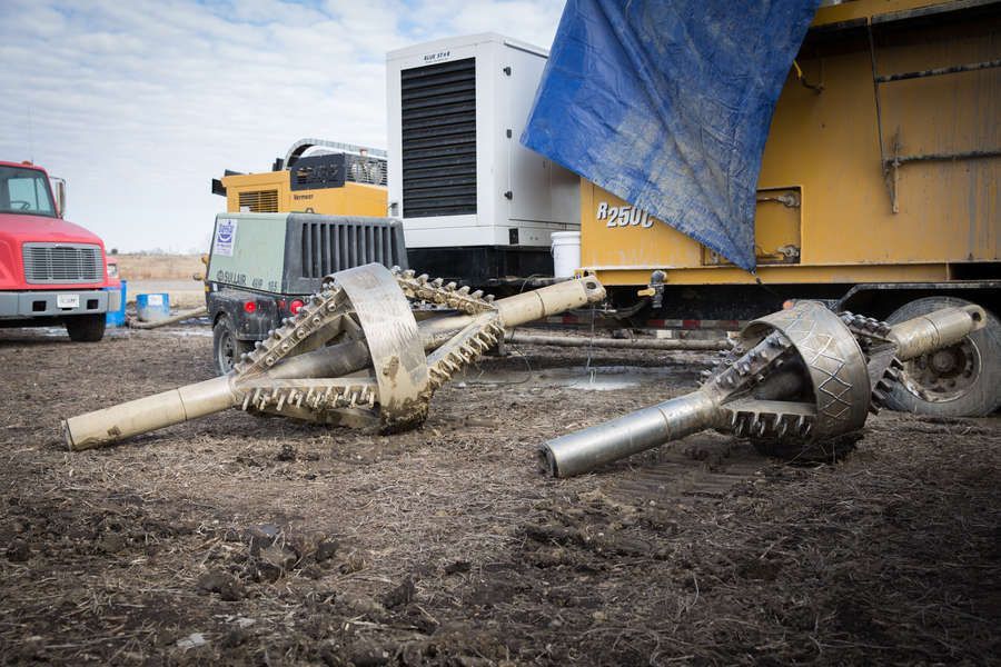 Two large drill bits on the ground at a construction site, with various vehicles and equipment visible in the background under a cloudy sky.