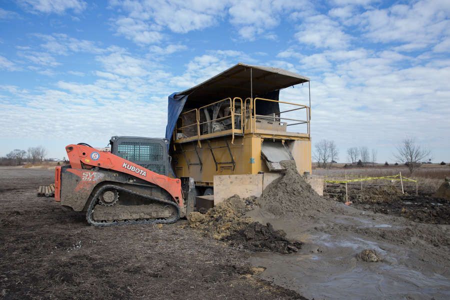 A skid steer loader dumping dirt into a large yellow soil screening machine in a field, under a blue sky.
