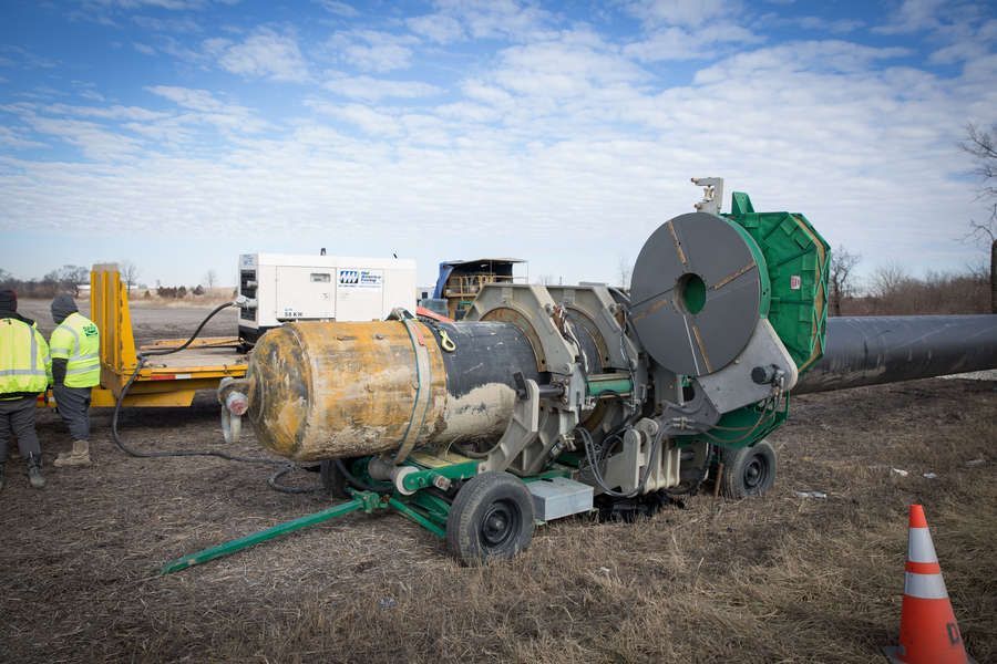 Large pipe welding machine in use outdoors, with workers present. Green, yellow, and orange equipment, pipe, and trailer, set in a field.