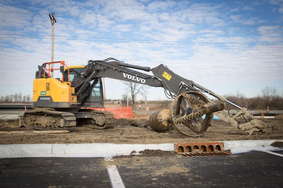 A yellow Volvo excavator with a rotary cutter digging into dirt.  A pipe section rests nearby on a construction site under a cloudy sky.