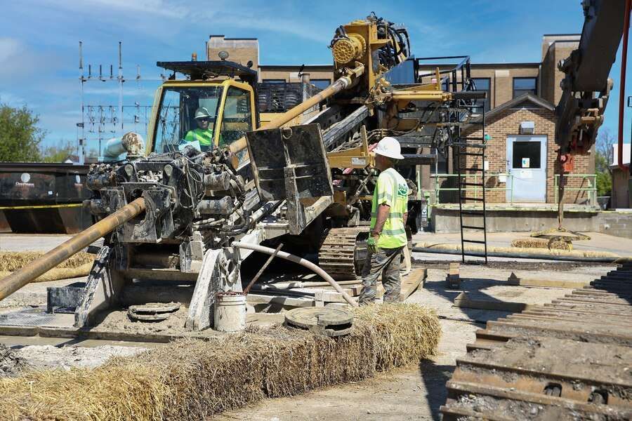 Construction workers operating machinery, likely drilling, near a building. One person is inspecting equipment, another is in the cab.
