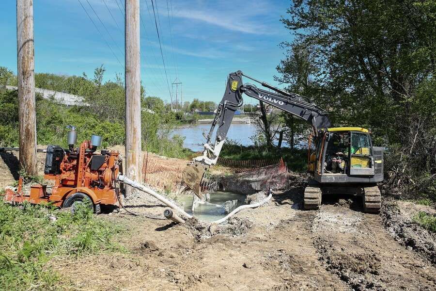 A hydraulic excavator and utility machine working near a power pole beside a body of water. The scene is outdoors on a sunny day.