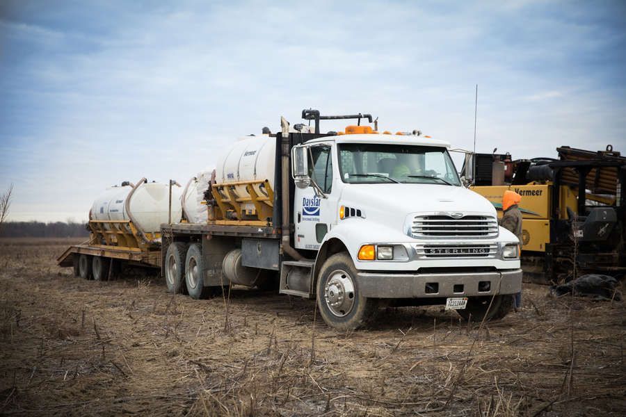 White tanker truck and trailer on a brown field under an overcast sky. The truck has a company logo on the door.