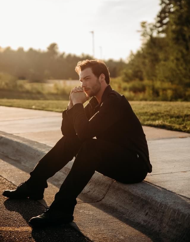 Man sits pensively on a curb, hands clasped. Golden sunlight bathes him; trees and road in the background.