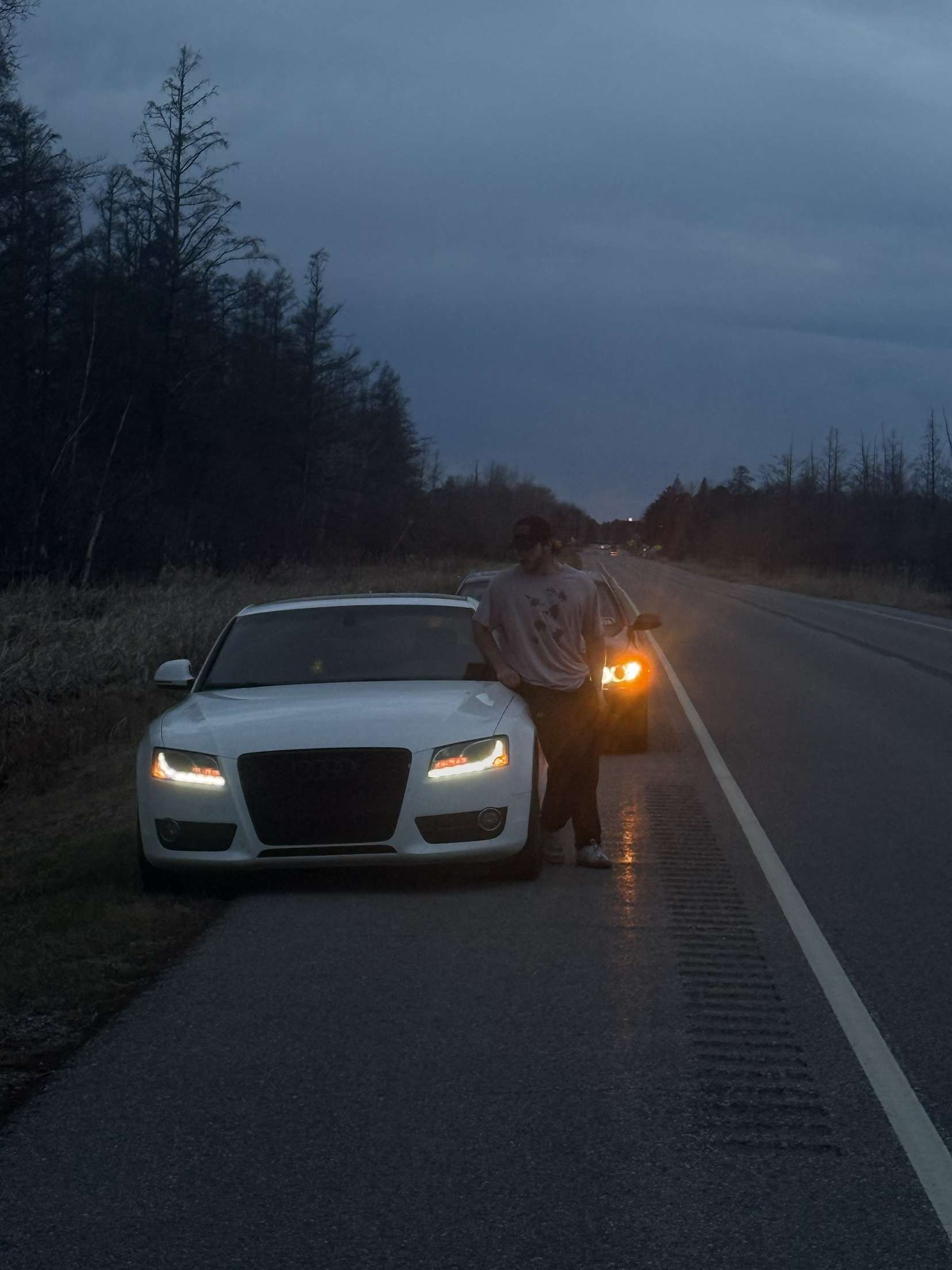 Person standing beside a white car on a road at dusk. Headlights are on, and trees are visible.