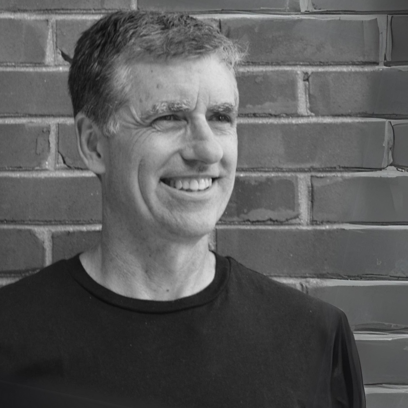 A man is smiling in front of a brick wall in a black and white photo.