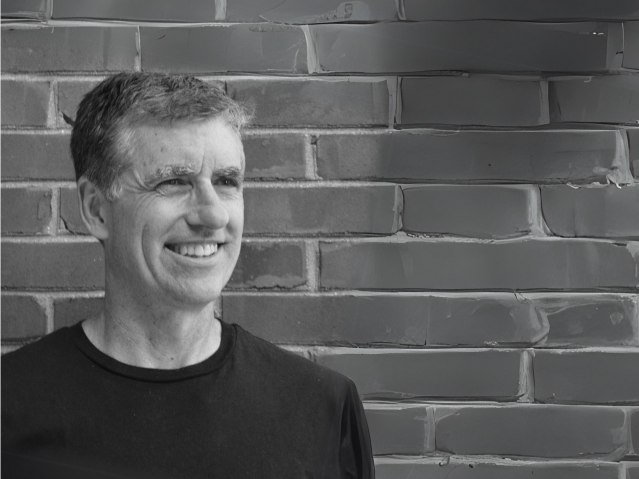 A man is smiling in front of a brick wall in a black and white photo.