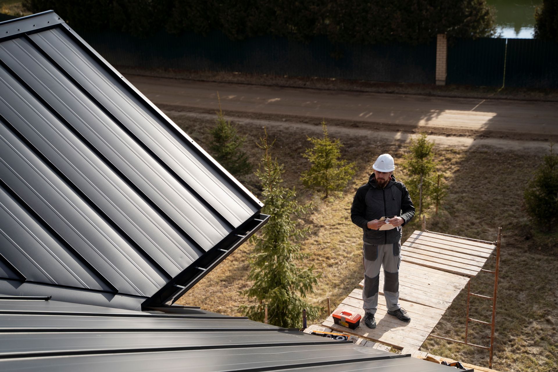 Roofer in white hardhat standing on a walkway, checking a device next to a dark metal roof.