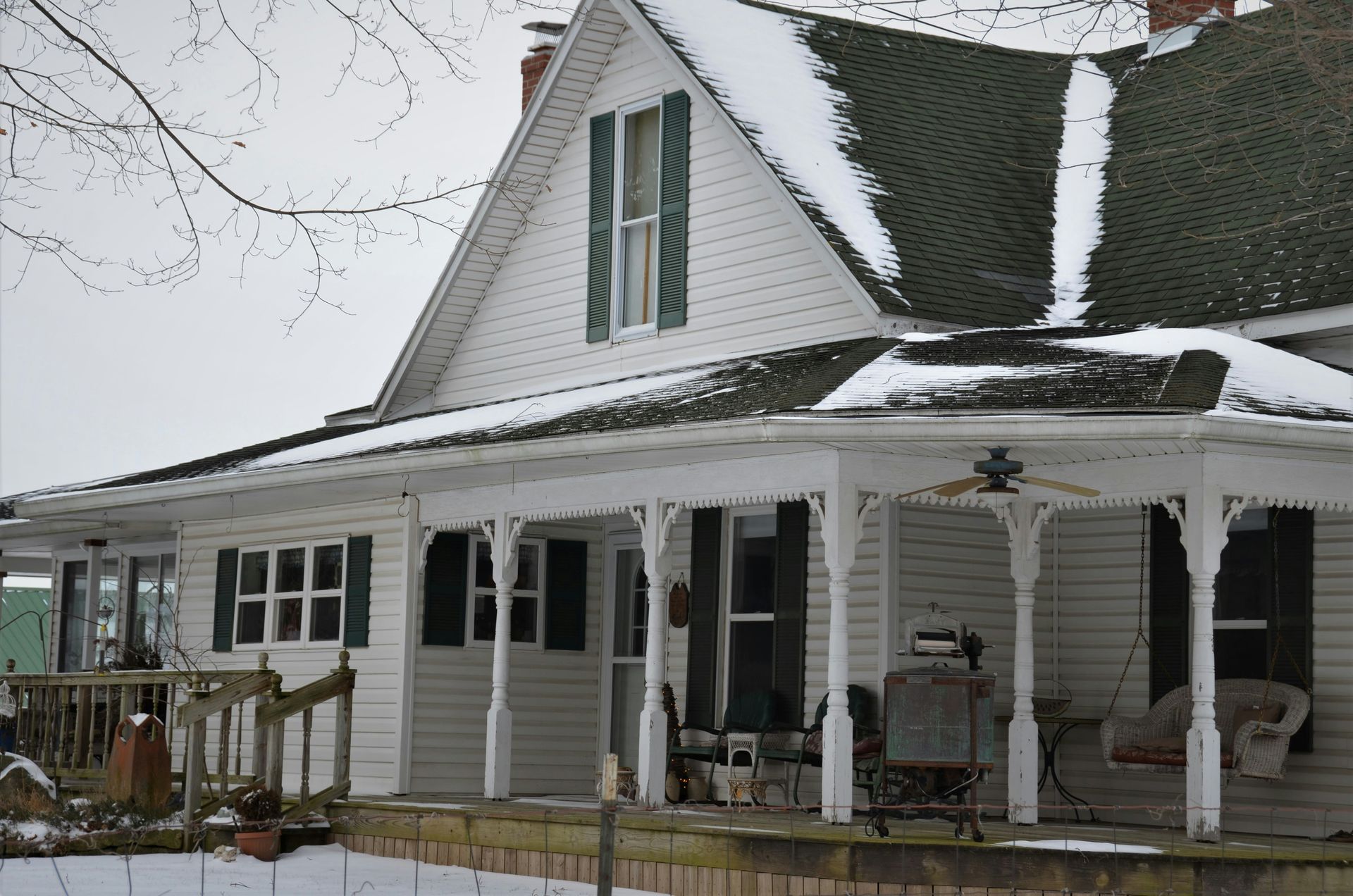 White house with snow-covered roof and porch. Green shutters, leafless trees, and overcast sky.