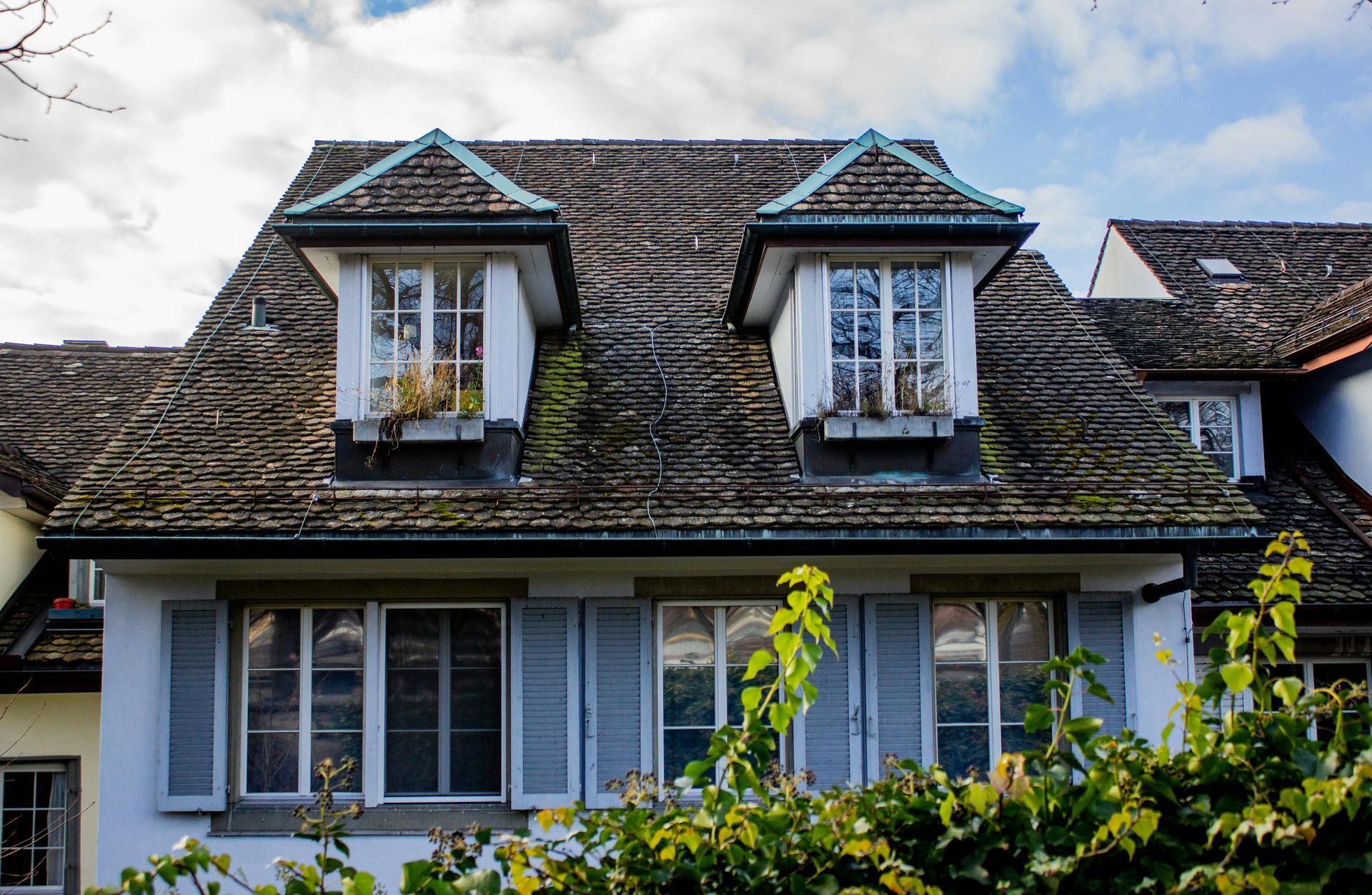 Stone house with mossy roof, dormer windows, and white shutters behind green foliage in Central PA
