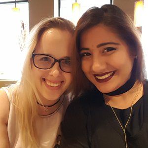 Two women are posing for a picture together and smiling for the camera . one of the women is wearing glasses and a choker.