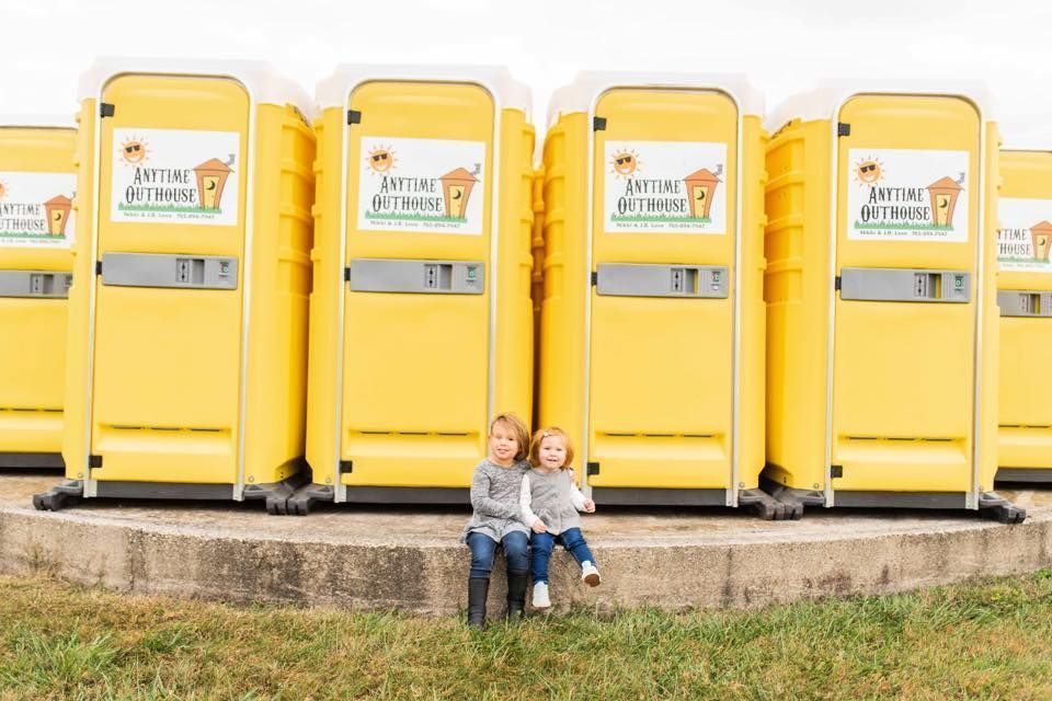 Two children sit in front of a row of yellow portable toilets with Anytime Outhouse logos