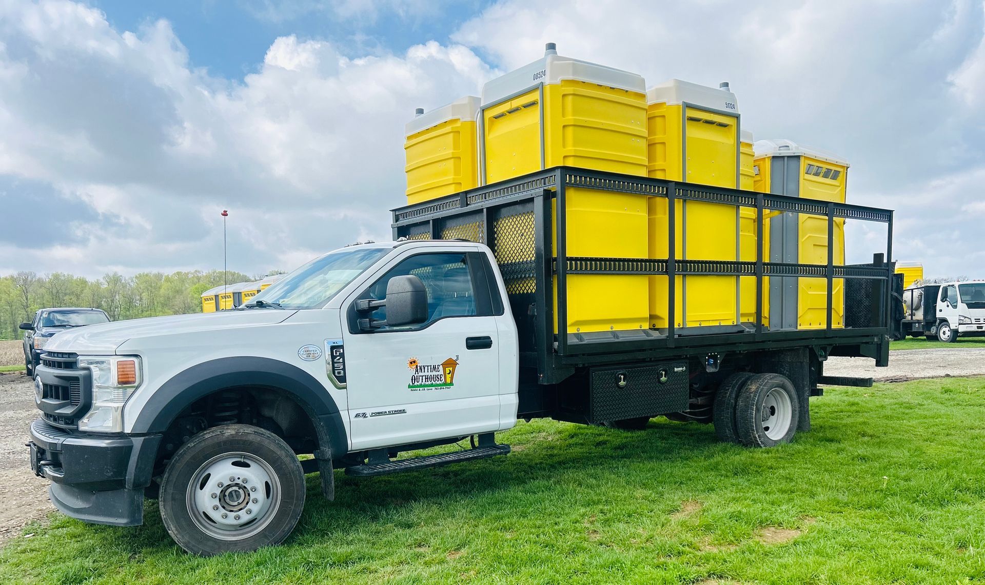 White truck loaded with yellow portable toilets on a grassy field under a cloudy sky.