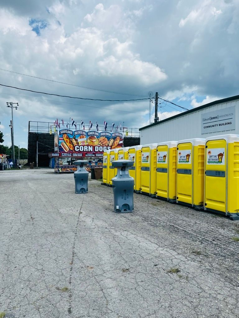 Yellow portable restrooms lined up outdoors, with handwashing stations. Cloudy sky and a fair in the background.