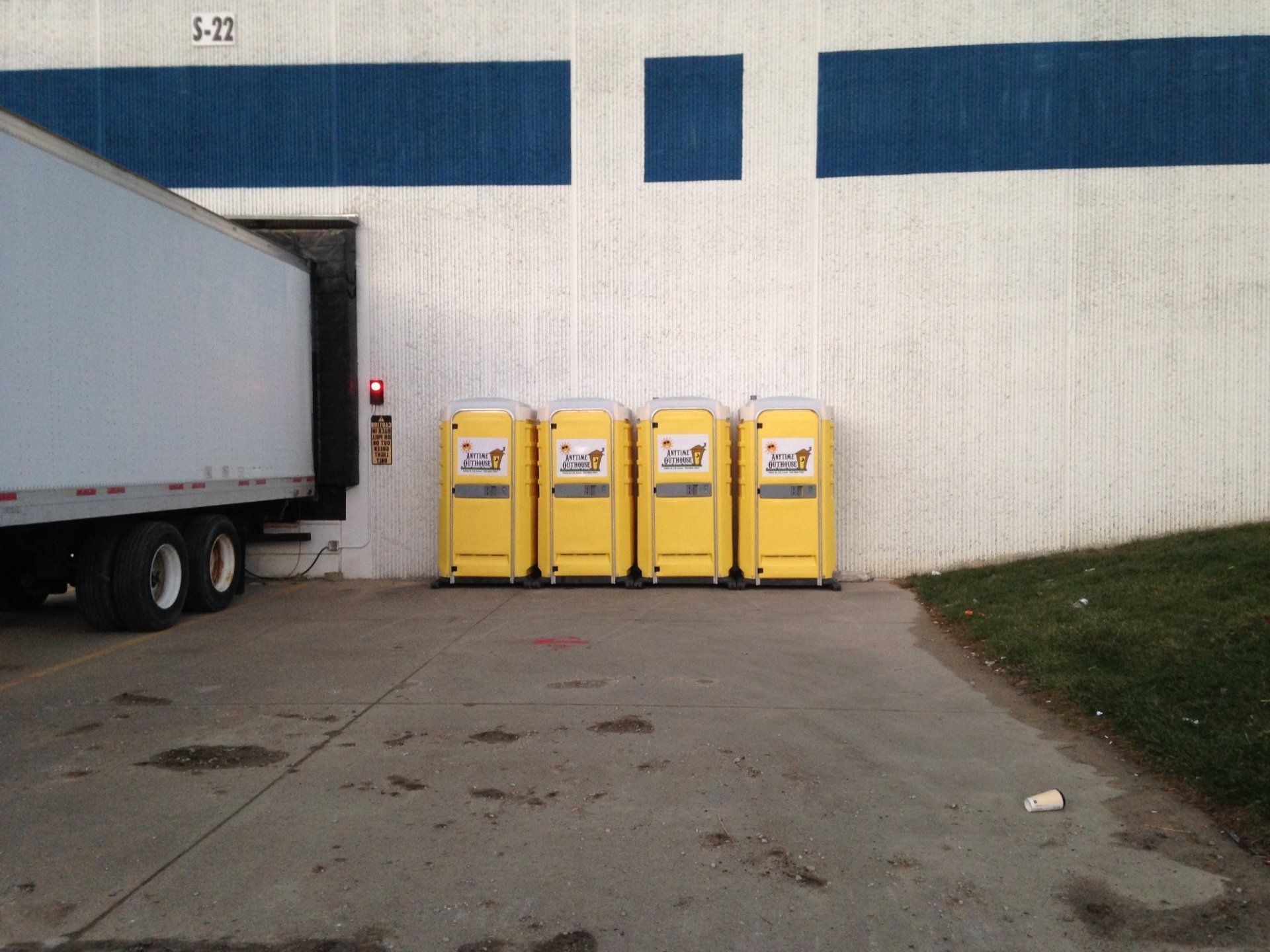 Four yellow portable toilets lined up in front of a warehouse with a truck backed up to a loading dock.