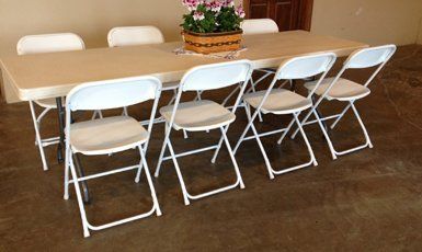 Long rectangular table with six white folding chairs; a flower arrangement in a basket sits in the center.