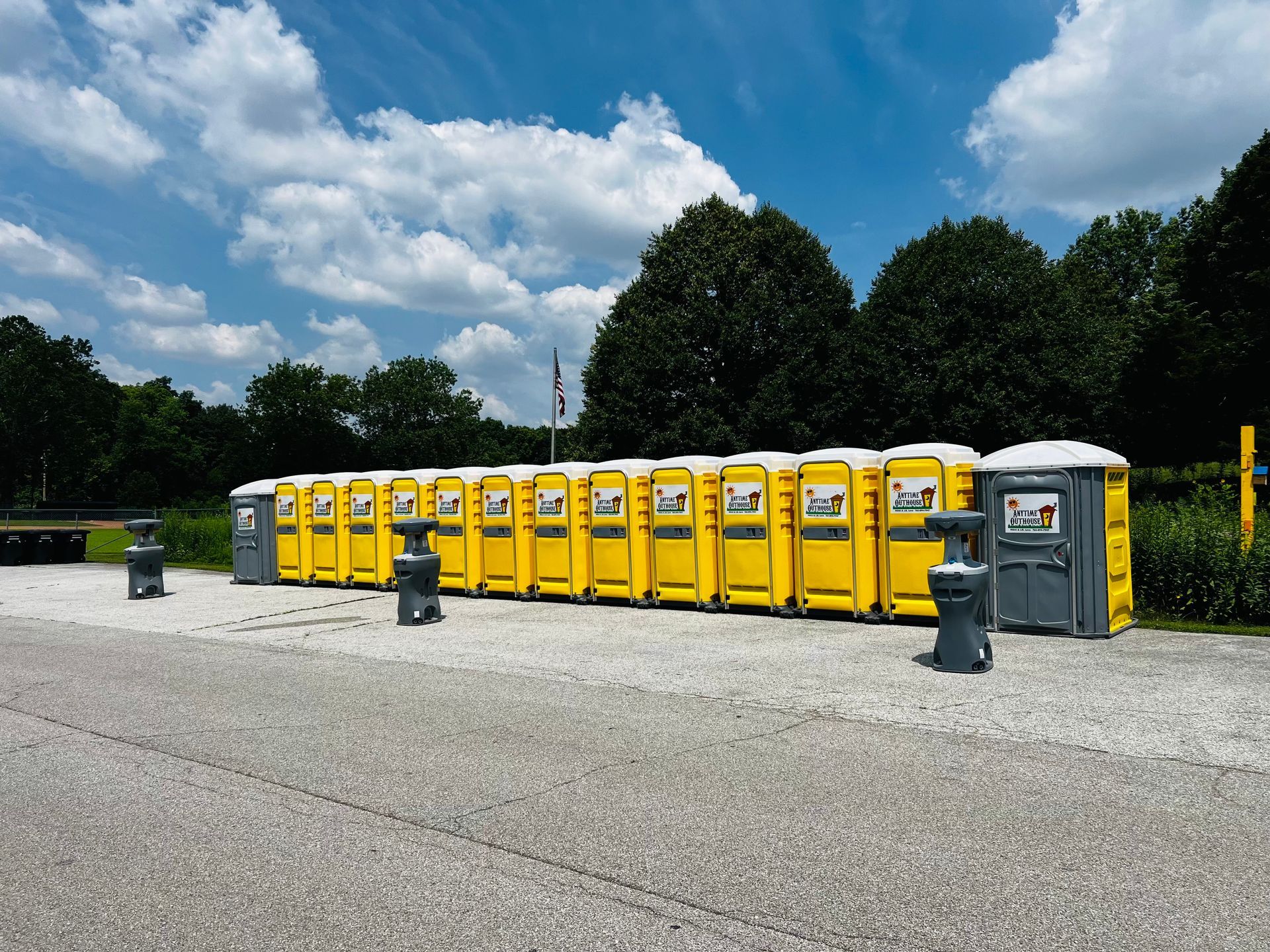 A row of yellow portable toilets, on a paved area, with trees and a cloudy blue sky in the background.