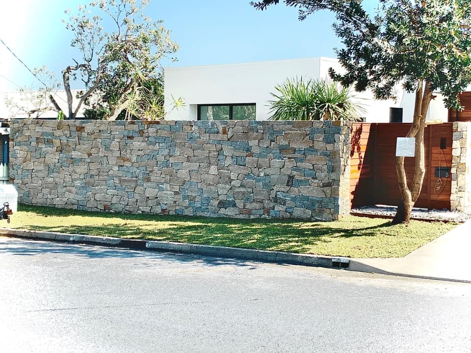 A Stone Wall Surrounds A Lush Green Yard In Front Of A House — Creative Landscapes Coffs Harbour In Korora, NSW
