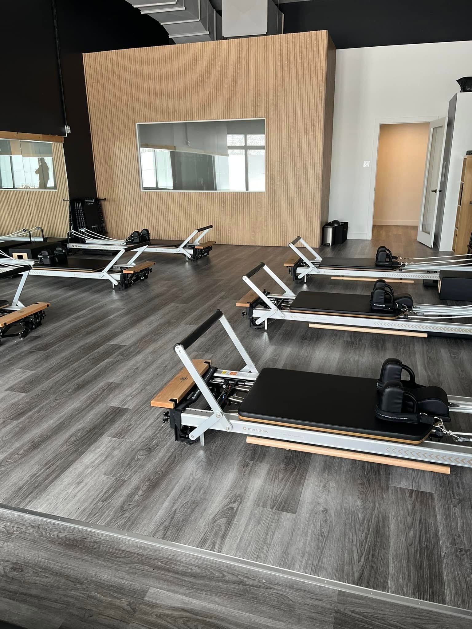 Studio de Pilates avec plusieurs appareils Reformer sur un sol imitation bois, contre un mur lambrissé, pièce lumineuse.