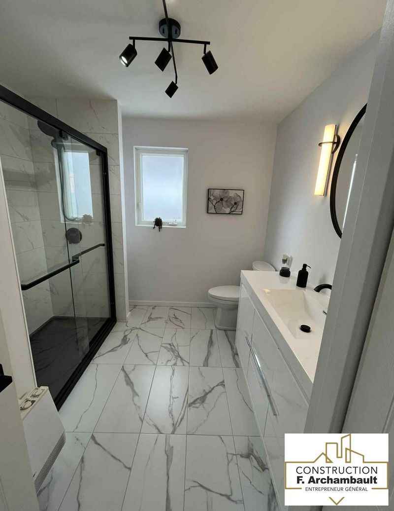 Salle de bains moderne blanche avec carrelage au sol imitation marbre, touches de noir et douche en verre.