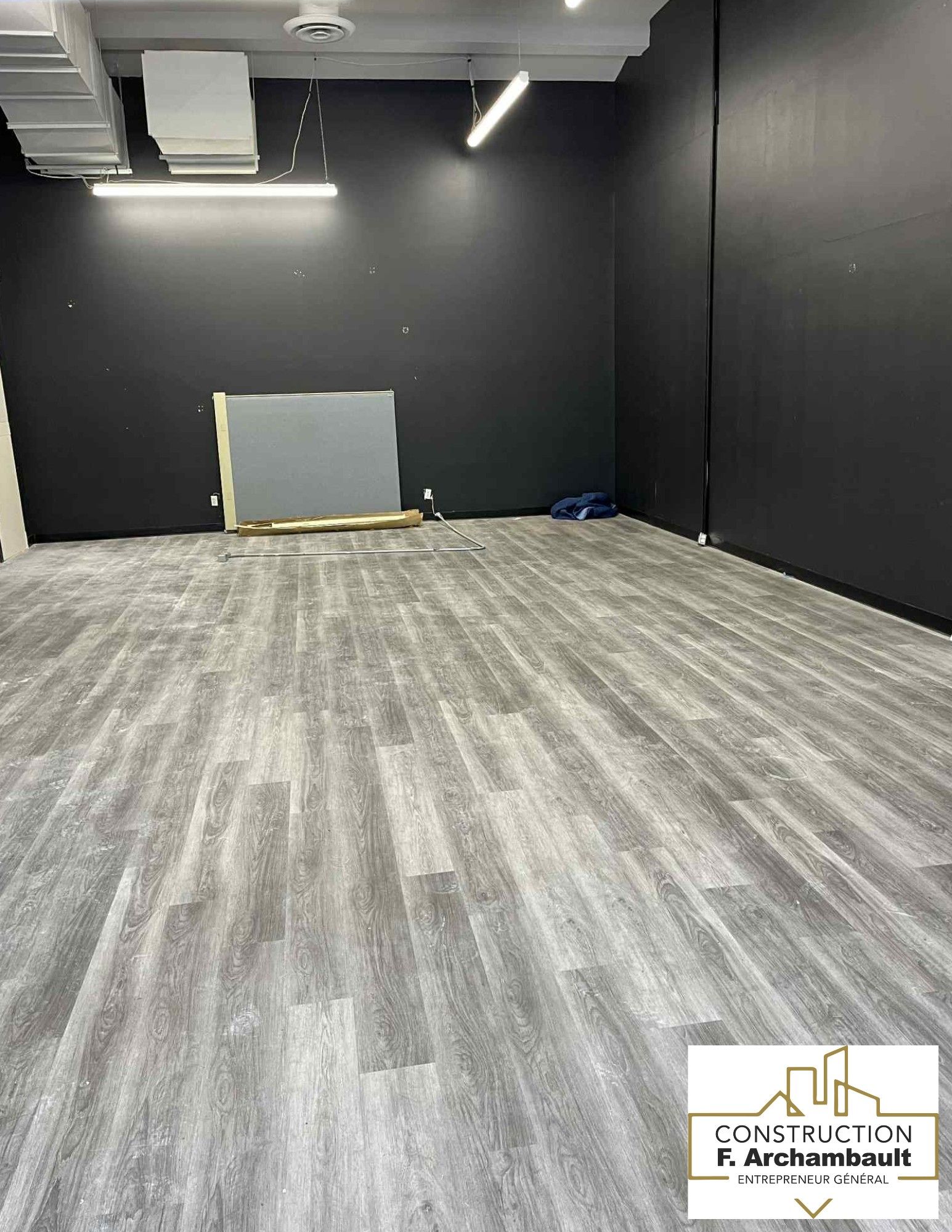 Pièce vide avec un sol imitation bois gris et des murs gris foncé, éclairée par des plafonniers.