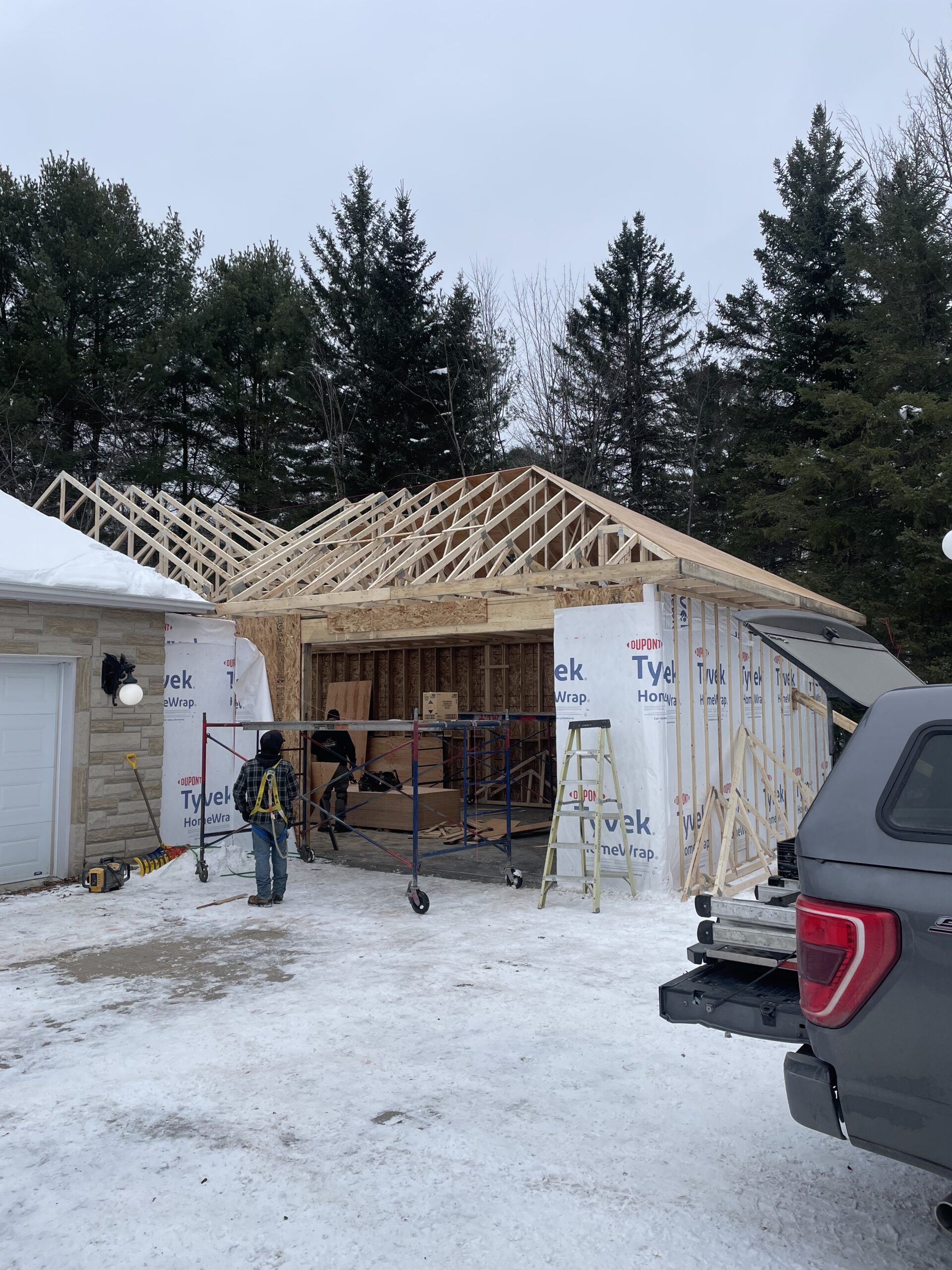 Garage en construction, charpente en bois, sol enneigé. Deux ouvriers, camion garé à proximité, arbres en arrière-plan.