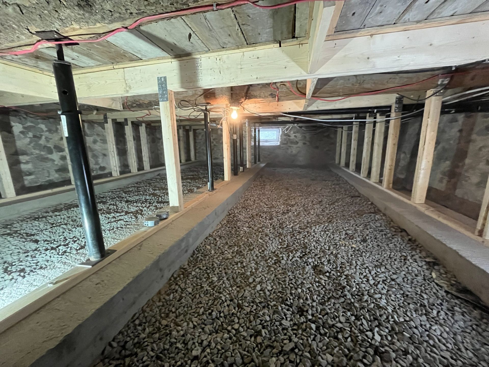 Vide sanitaire recouvert de gravier avec poutres en bois, poteaux de soutien et vérins réglables.