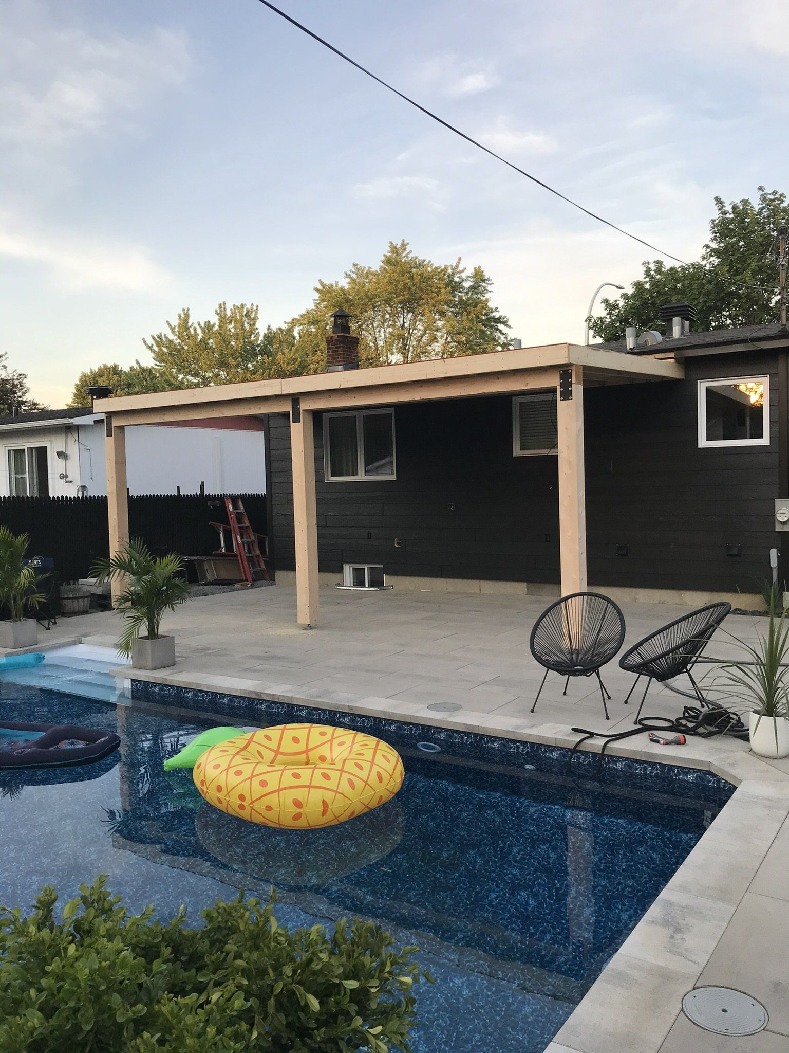 Au bord de la piscine, terrasse couverte, bouée ananas, façade sombre de la maison. Piscine bleue, poutres de soutien en bois clair.