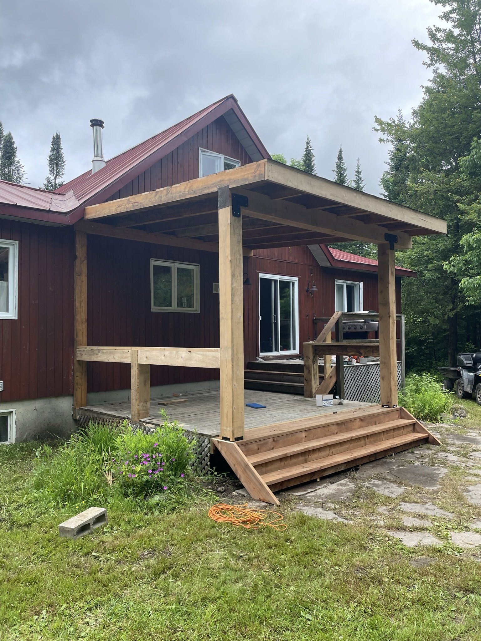 Terrasse en bois et pergola attenantes à une maison rouge, marches descendant vers une cour gazonnée, journée nuageuse.