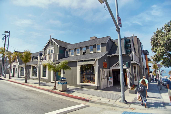A gray, multi-story building with a peaked roof stands on a sunny street corner with palm trees and pedestrians.