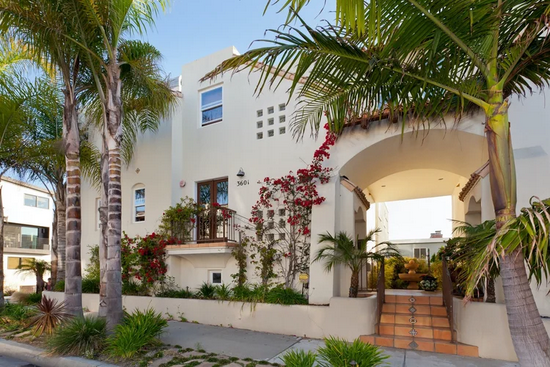 A two-story, white stucco building with a large arched entryway, tiled steps, and palm trees in a sunny, coastal setting.