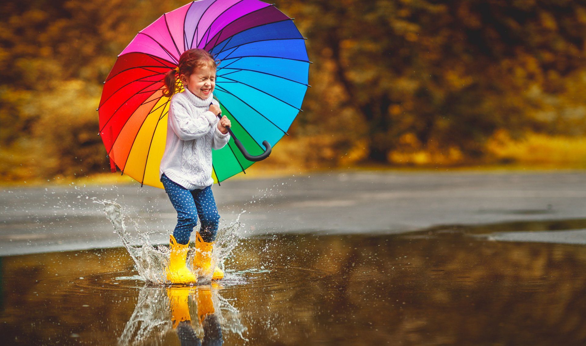 Smiling girl with colorful umbrella splashing in a puddle.