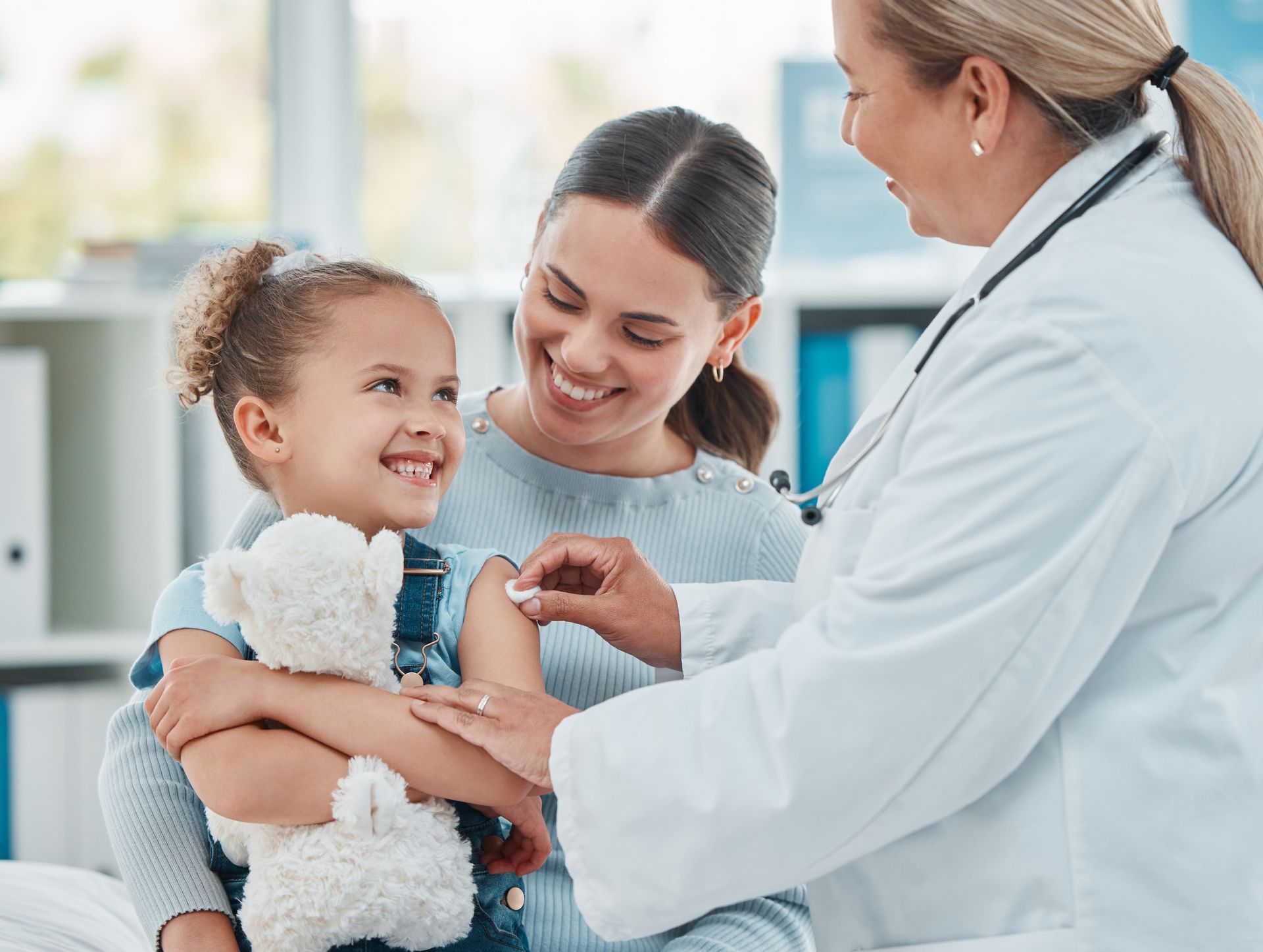 A little girl is getting an injection from a doctor while holding a teddy bear.