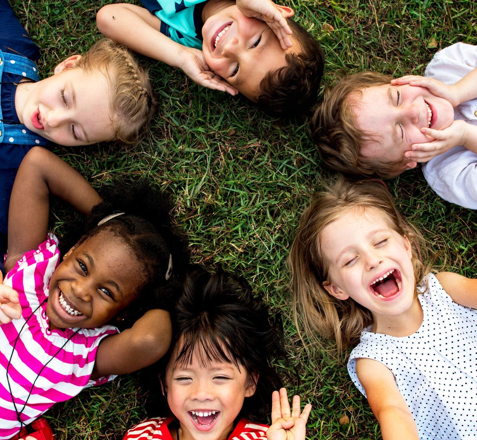 A group of smiling children are laying in the grass in a circle.