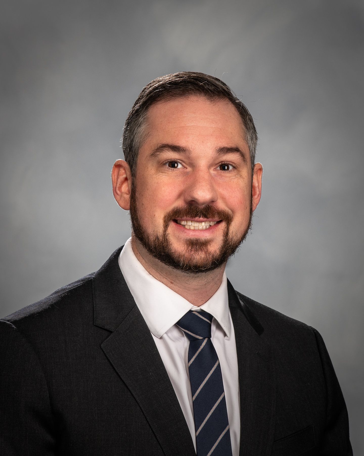 Man in a blue suit and tie, smiling, outdoors with blurred greenery in the background.