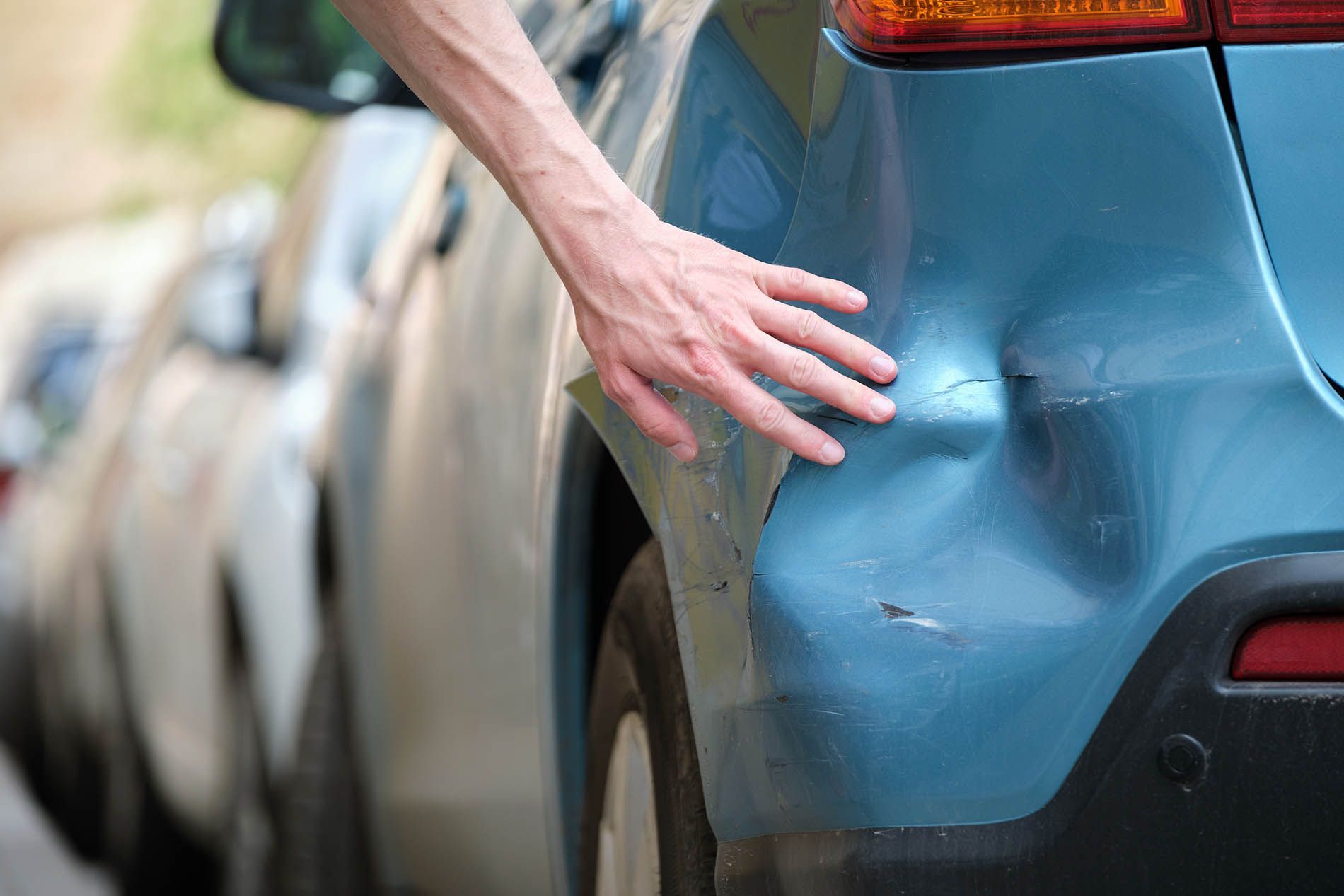 Person's hand touching a dented, blue car bumper, likely after a collision, outdoors.