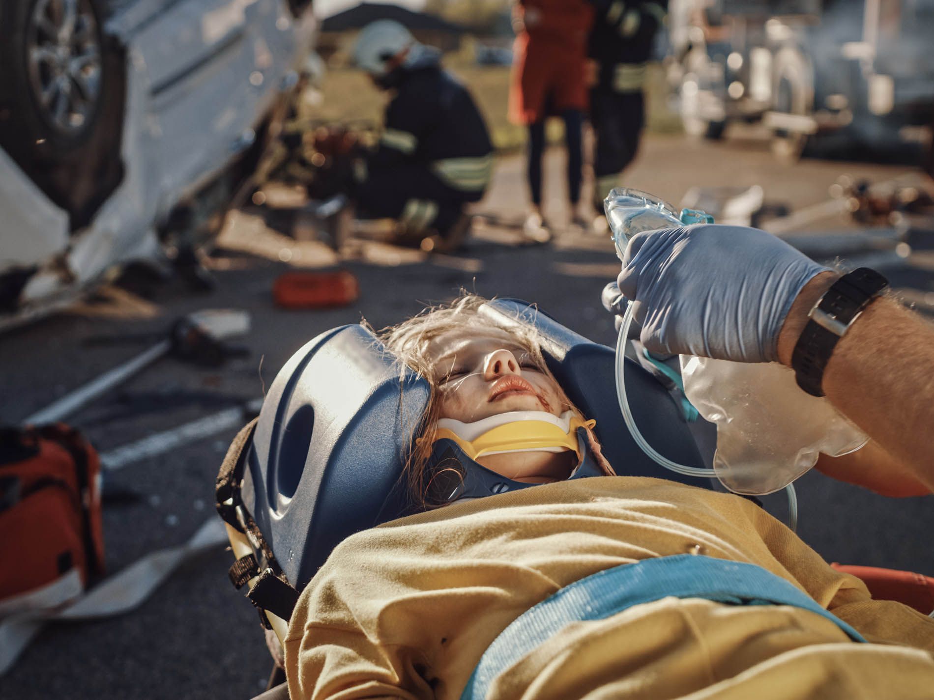 Child injured in car accident, receiving oxygen with medical personnel and debris in background.
