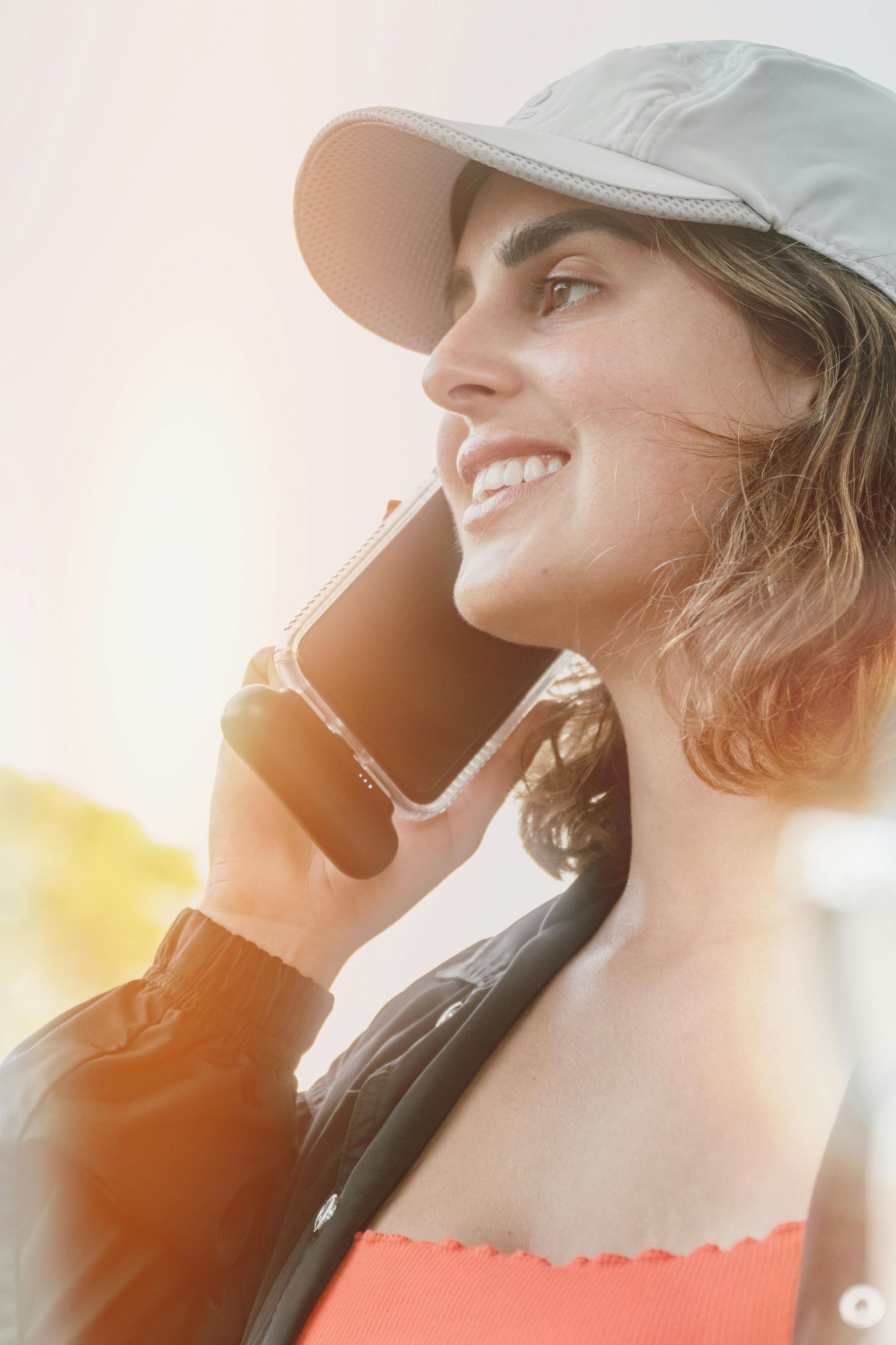 Woman in cap smiles while talking on phone, outside. Orange top, black jacket, sunny.